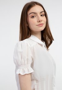 Weißes Blusenshirt mit kurzen Puffärmeln, Knopfverschluss vorne und gerafften Details an den Ärmelbündchen. Strukturierter Stoff.