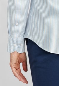 Camicia a maniche lunghe a righe blu chiaro e bianche con polsini abbottonati, caratterizzata da un orlo arrotondato e una texture di tessuto liscia.