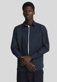 Camicia blu navy con maniche lunghe, colletto classico e un sottile dettaglio del logo sul petto. Tessuto liscio, vestibilità strutturata.