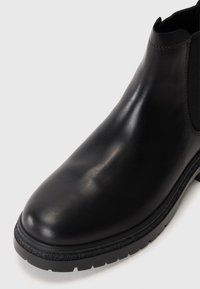 Bocanci scurti din piele neagră, textură netedă, bot rotund, panouri laterale elastice și o talpă groasă din cauciuc cu modele de tracțiune.