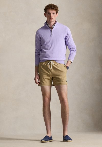 Νέος άνδρας που φοράει πουλόβερ με φερμουάρ στο στήθος σε λιλά χρώμα, μπεζ σορτς με κορδόνια, ναυτικά espadrilles και ρολόι χειρός, στέκεται μπροστά σε ουδέτερο φόντο.