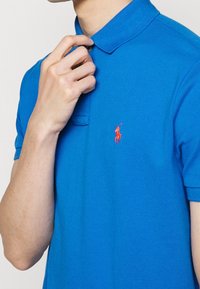 Niebieska koszulka polo wykonana z teksturowanej tkaniny, z kołnierzykiem i krótkimi rękawami. Na lewej piersi znajduje się małe, czerwone, haftowane logo.