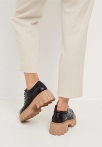 Chaussures à plateforme en cuir noir avec une semelle épaisse beige, présentant un bout arrondi, des lacets et une texture lisse. Portées avec un pantalon de couleur claire.