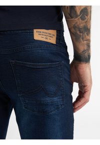 Tmavě modré džíny z denimu s koženou nášivkou na pase. Mezi charakteristiky patří prošívané zadní kapsy a slim fit střih.