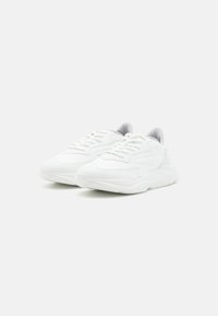 White athletic shoes featuring a textured fabric upper, padded tongue, and thick sole. Laces in front, slight curvature in design.