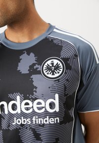 Camiseta deportiva transpirable en gris y negro con un patrón digital, un cuello redondo y un logo de un águila con una corona. "Indeed" impreso de manera prominente.