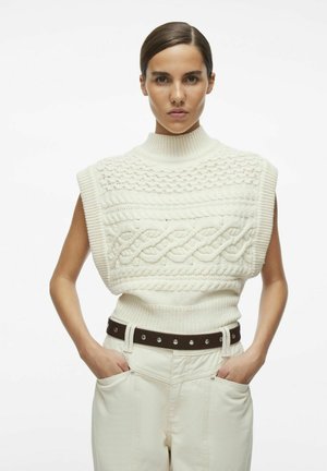 Maglione senza maniche a collo alto bianco con motivi a treccia, abbinato a pantaloni chiari a vita alta e una cintura scura.