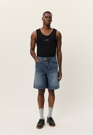 Muž v čiernom tielku s nápisom, modrých džínsových krátkych nohaviciach, sivých ponožkách a čiernych topánkach stojaci pred jednoduchým pozadím.