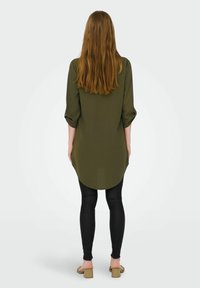 Olivegrünes langes Hemd mit aufgekrempelten Ärmeln und abgerundetem Saum, kombiniert mit schwarzen Leggings und beigefarbenen Sandalen mit Blockabsatz, von hinten gesehen.