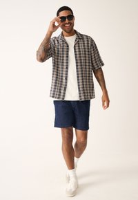 Chemise à manches courtes à carreaux bleu marine et blanc, t-shirt blanc en dessous, associée à un short bleu marine et des baskets blanches, accessoirisée avec des lunettes de soleil.