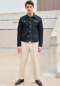 Dark denim jacket with silver buttons, paired with light beige trousers and black loafers, set against a neutral-toned background.