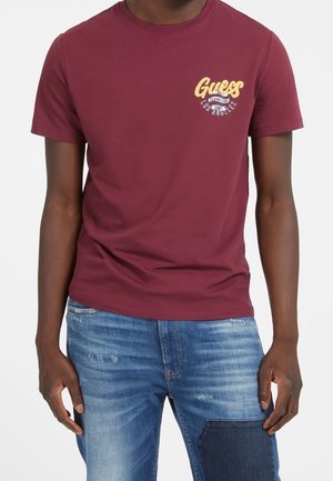 Homme portant un t-shirt Guess bordeaux avec un logo jaune sur la poitrine gauche et un jean en denim bleu avec une tache foncée sur la cuisse droite.