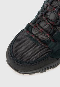 Dunkelgrün und schwarze Sneaker aus Netz- und Wildledermaterial, strukturierte Oberfläche, rote Nähte und eine Gummisohle mit Grip-Mustern.