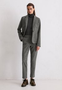 Grijs gestructureerd wollen pak, bestaande uit een op maat gemaakte blazer en aansluitende pantalon, gecombineerd met een donkere coltrui en bruine veterschoenen.