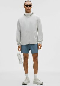 Vaaleanharmaa vetoketjulla varustettu huppari, yhdistettynä sinisiin shortseihin. Malli pitää kädessään valkoista vesipulloa, ja hänellä on harmaat lenkkarit ja aurinkolasit.