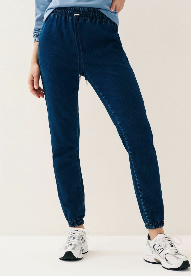 Pantalon de jogging bleu marine avec taille et poignets élastiques porté avec des chaussures de sport blanches sur une personne debout.