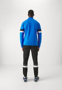 Blauwe sporttop met lange mouwen en zwart-witte strepen op de mouwen, gecombineerd met zwarte broeken met witte banden bij de boorden en blauwe schoenen.