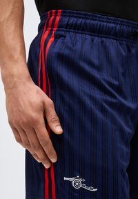 Roka počiva ob temno modrih športnih kratkih hlačah z rdečimi črtami in belim logotipom topa blizu žepa.