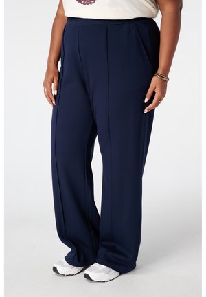 Personne portant un pantalon bleu marine ample avec des coutures à l'avant, associé à des baskets blanches et un haut clair, debout devant un fond uni.