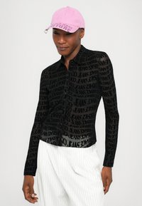 Black sheer button-up shirt with repeated text pattern. Paired with a pink baseball cap. White pinstriped pants visible.
