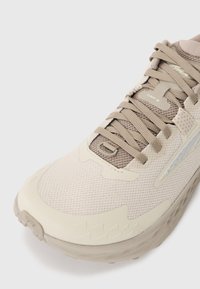 Chaussure de trail en beige et taupe avec tige en mesh, bout renforcé et semelle épaisse et texturée, étiquetée "TIMP 5" près des lacets.