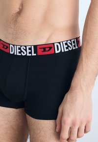 Boxer-briefs noirs avec une ceinture de marque portant le mot "DIESEL" en gras blancs et rouges. Le tissu semble lisse et élastique.