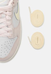 Deeltelijke weergave van een witte en lichtroze sneaker met zachte roze veters, naast twee opgerolde beige vervangende veters op een witte achtergrond.
