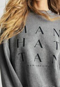 Grijze sweatshirt met zwarte tekst die "MANHATTAN" en "NEW YORK CITY" spelt, met een klassieke ronde hals en een zachte textuur.