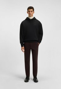 Felpa nera con cappuccio e vestibilità comoda, con logo sul petto, abbinata a pantaloni bordeaux scuro e scarpe nere con lacci.