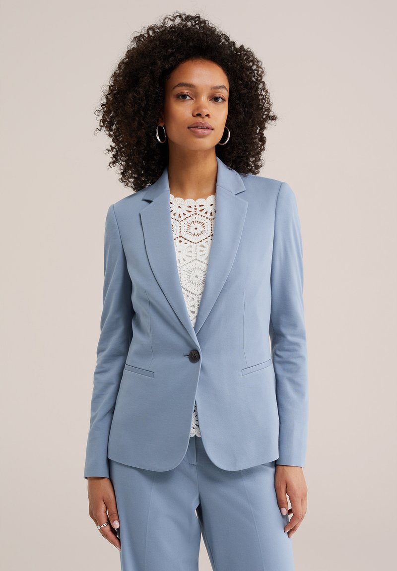 Femme aux cheveux bouclés portant une veste et un pantalon de costume bleu clair sur un haut blanc en crochet, debout devant un fond beige.
