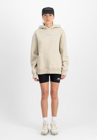 Beige oversized hoodie met een voorkruis, blauwe tekst 'STUDIO EDITION'. Zwarte fietsbroek en lichtgekleurde sneakers met zichtbare veters.