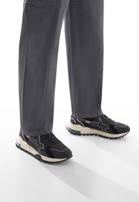 Schwarze Sportschuhe aus strukturiertem Material, mit einer cremefarbenen Sohle und gummierten Traktionsnoppen. Kombiniert mit locker sitzenden grauen Hosen.