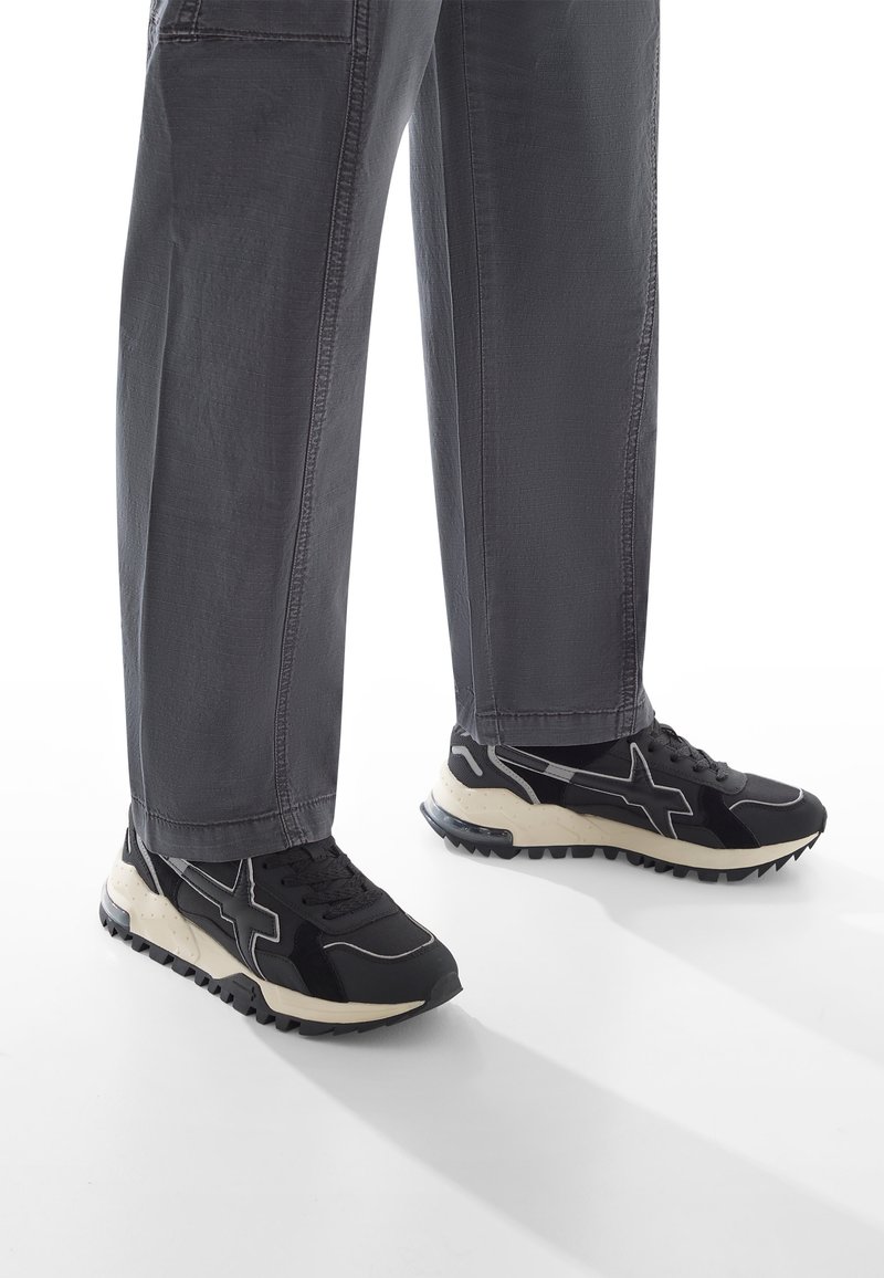 Schwarze Sportschuhe aus strukturiertem Material, mit einer cremefarbenen Sohle und gummierten Traktionsnoppen. Kombiniert mit locker sitzenden grauen Hosen.