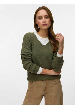 Ženska z dolgimi rjavimi lasmi, oblečena v olivno zelen pulover z V-izrezom čez belo srajco in svetlo rjave hlače, stoji z rokami prekrižanimi.