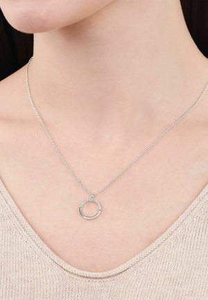 Zilveren ketting met een kleine open cirkel hanger, gedeeltelijk versierd met kleine sprankelende steentjes, gedragen om de nek van een persoon.