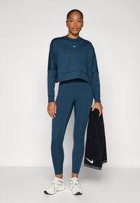 Sweatshirt court bleu marine avec des poignets côtelés, associé à un legging assorti. Le modèle tient une serviette noire avec un logo Nike blanc. Des baskets blanches complètent le look.