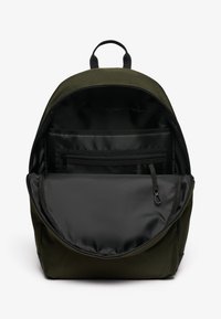 Mochila verde oscura con el compartimento principal abierto, mostrando el forro interior negro, un bolsillo de malla con cremallera y un clip para llaves en el interior.