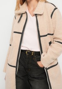 Femme portant un manteau sherpa beige avec bordure noire sur un pull rose pâle à col en V et un jean noir avec ceinture noire et boucle dorée, main dans la poche.