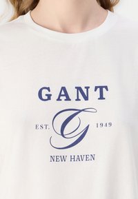 T-shirt in cotone bianco con grafica blu navy che presenta il testo "GANT EST. 1949 NEW HAVEN" e un design stilizzato della lettera "G".