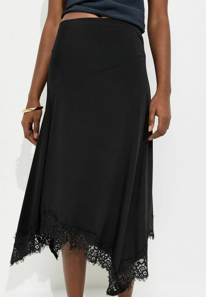 Woman wearing a black asymmetrical midi skirt with lace trim along the hem, paired with a dark top and a gold bangle on her wrist.