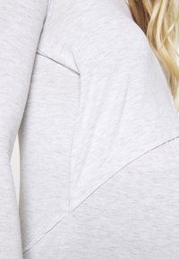 Top de tecido cinza claro com um design texturizado, costura diagonal e ajuste solto. Mangas longas e material macio e elástico.