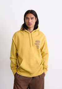 Gelber Hoodie mit einer Fronttasche und Grafikdruck auf der Brust. Der Stoff wirkt weich und hat eine lockere Passform. Kombiniert mit braunen karierten Hosen.