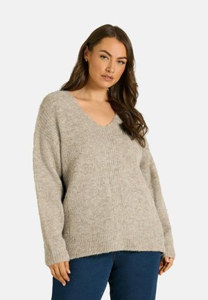 Femme aux longs cheveux bruns portant un pull beige ample à col en V et un jean bleu foncé sur un fond clair uni.