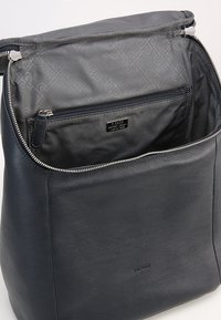 Intérieur d'un sac en cuir noir présentant une texture lisse, une doublure grise et une poche zippée intérieure affichant une plaque de logo.