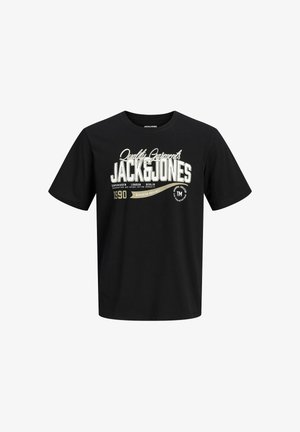 Teenager med krøllet hår iført en grå hættetrøje med lynlås, sort grafisk t-shirt, brede blå jeans og sorte sneakers, står og smiler.