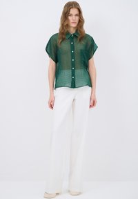 Camicia verde trasparente a maniche corte con un motivo testurizzato, abbinata a pantaloni bianchi a gamba larga e scarpe di colore chiaro.