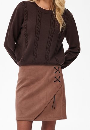Femme portant un pull en maille texturée marron foncé et une jupe portefeuille marron clair avec un détail en lacets noirs sur le côté.