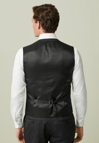 Schwarze Satinweste mit glatter Textur, verstellbarem Riemen auf der Rückseite, kombiniert mit einem weißen Hemd. Von hinten gezeigt.