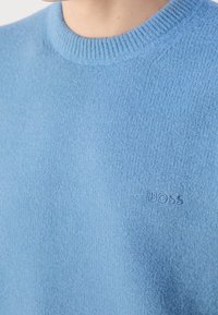 Ljusblå stickad tröja med en texturerad yta, med en ribbad rund halsringning och en subtil präglad logotyp på bröstet.