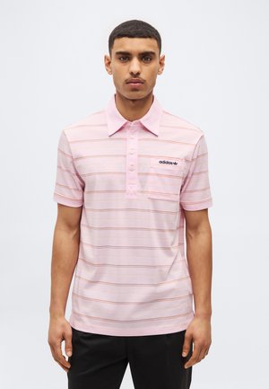 Vyras, dėvintis šviesiai rožinę trumpomis rankovėmis Adidas polo marškinėlį su plonomis horizontaliomis juostelėmis ir krūtinės kišene, stovintis priešais vienspalvį foną.